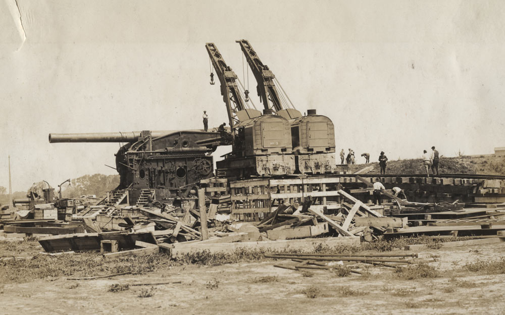 Sephia photograph of a construction site. Two cranes and scaffolding surround a house-sized gun.