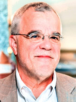 Headshot of a middle-aged white man with grey hair and stubble wearing glasses and a tan suit jacket