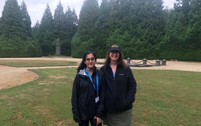 Photograph of two female high school students wearing black windbreaker jackets standing on a grass lawn in a clearing. There is a small statue on a plinth behind them.