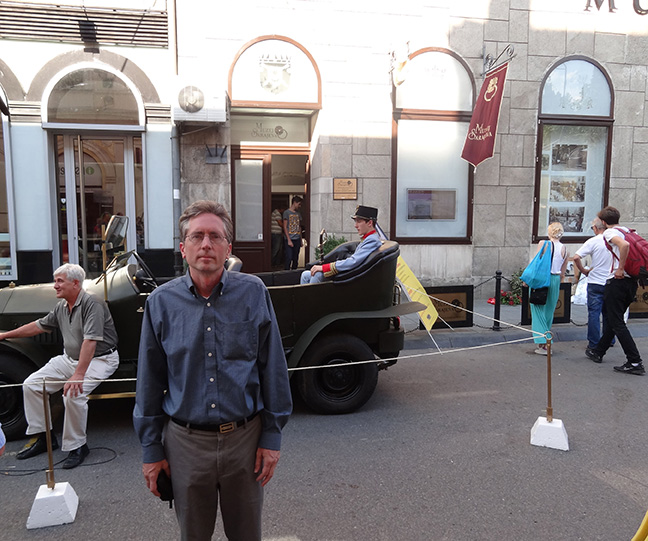 A middle-aged man standing in front of a model of an early 1900s automobile in a city street