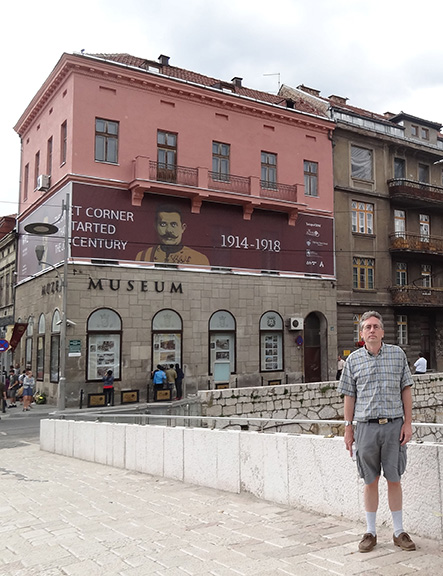 A white middle-aged man in a polo and cargo shorts standing in front of a corner building with a banner wrapped around two sides