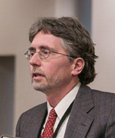 Headshot of a middle-aged white man with greying hair and stubble wearing glasses and a grey suit jacket