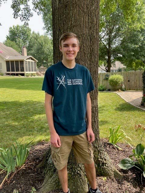 A young white man wearing a National WWI Museum and Memorial shirt posing in front of a tree