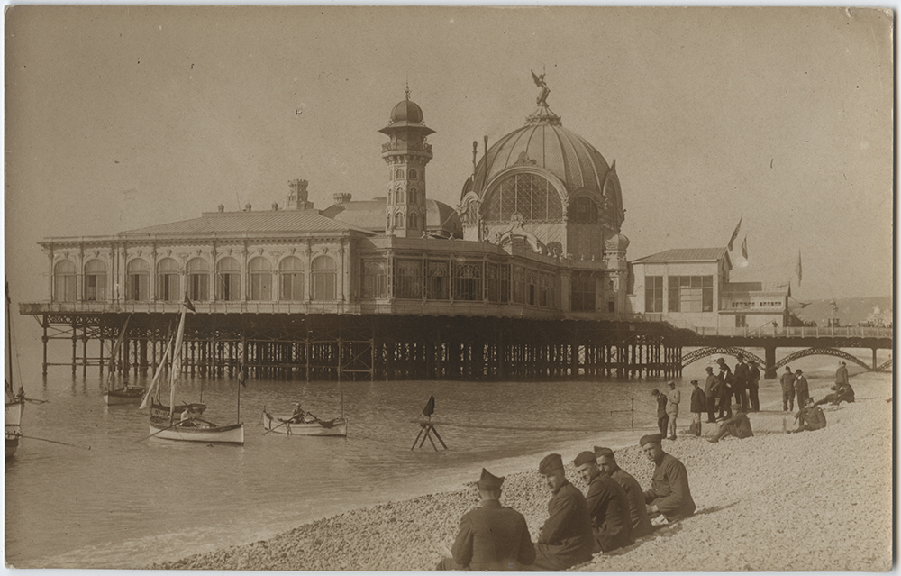 Sepia photograph of a beach in the foreground with soldiers and other beachgoers sitting on the sand. In the water are small rowboats and sailboats. In the background is a palace that extends out into the water on elevated pilings.