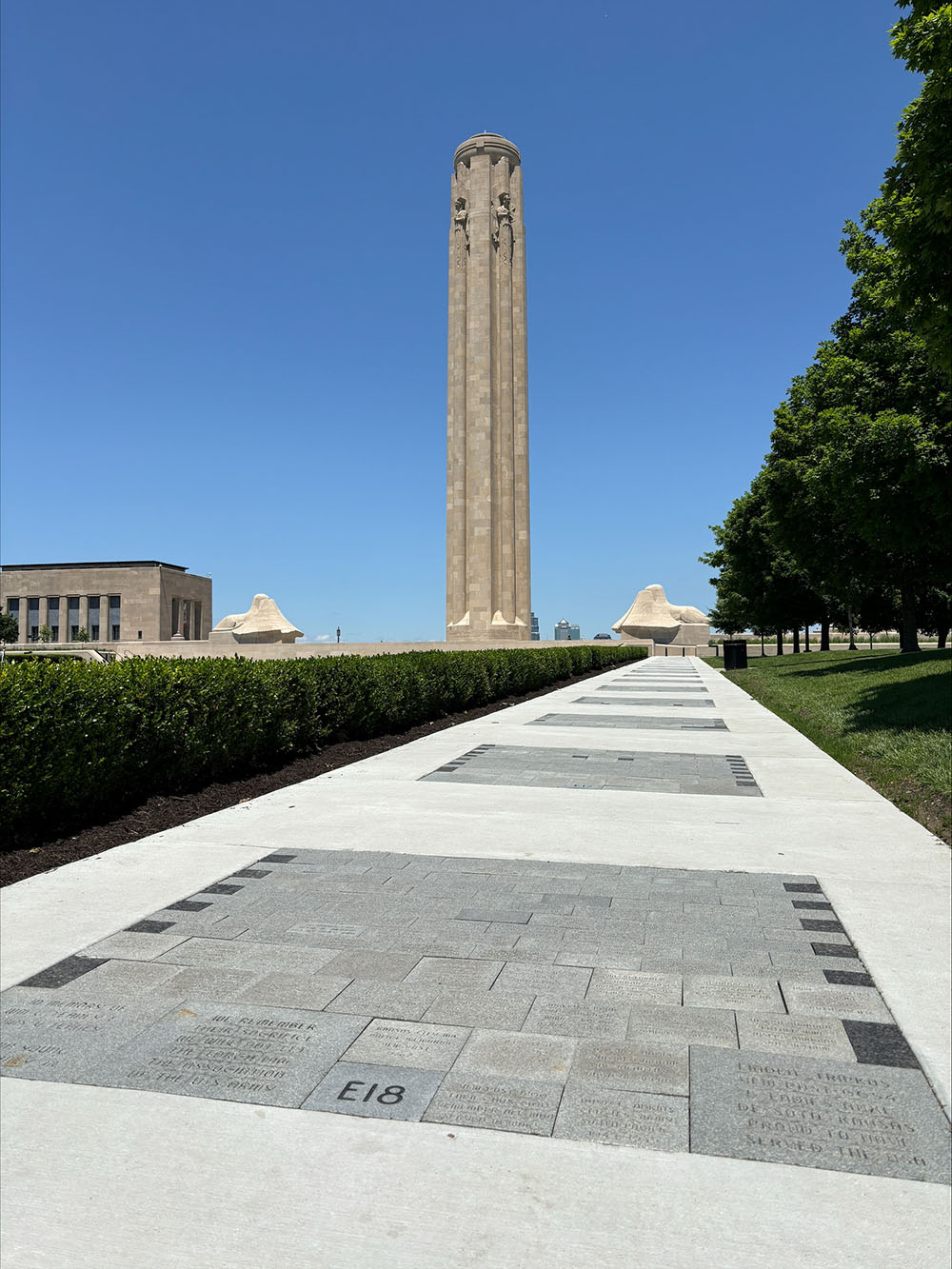 Modern photo of a wide sidewalk bordering a large lawn and shrubbery stretching away toward Memorial Courtyard and Liberty Memorial Tower in the distance. Engraved dark granite bricks are embedded periodically in the sidewalk.