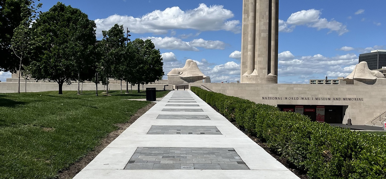 Modern photograph of a wide clean sidewalk bordering a large lawn and shrubbery, stretching towards Liberty Memorial Tower in the distance. Dark granite bricks are embedded at regular intervals in the sidewalk.