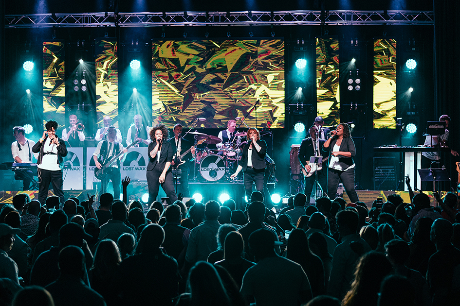 Modern photo of a stage in a darkened theater. The stage is illuminated with bright stage lights in white and blue. A large group of musicians and singers play energetically to an enthusiastic crowd.