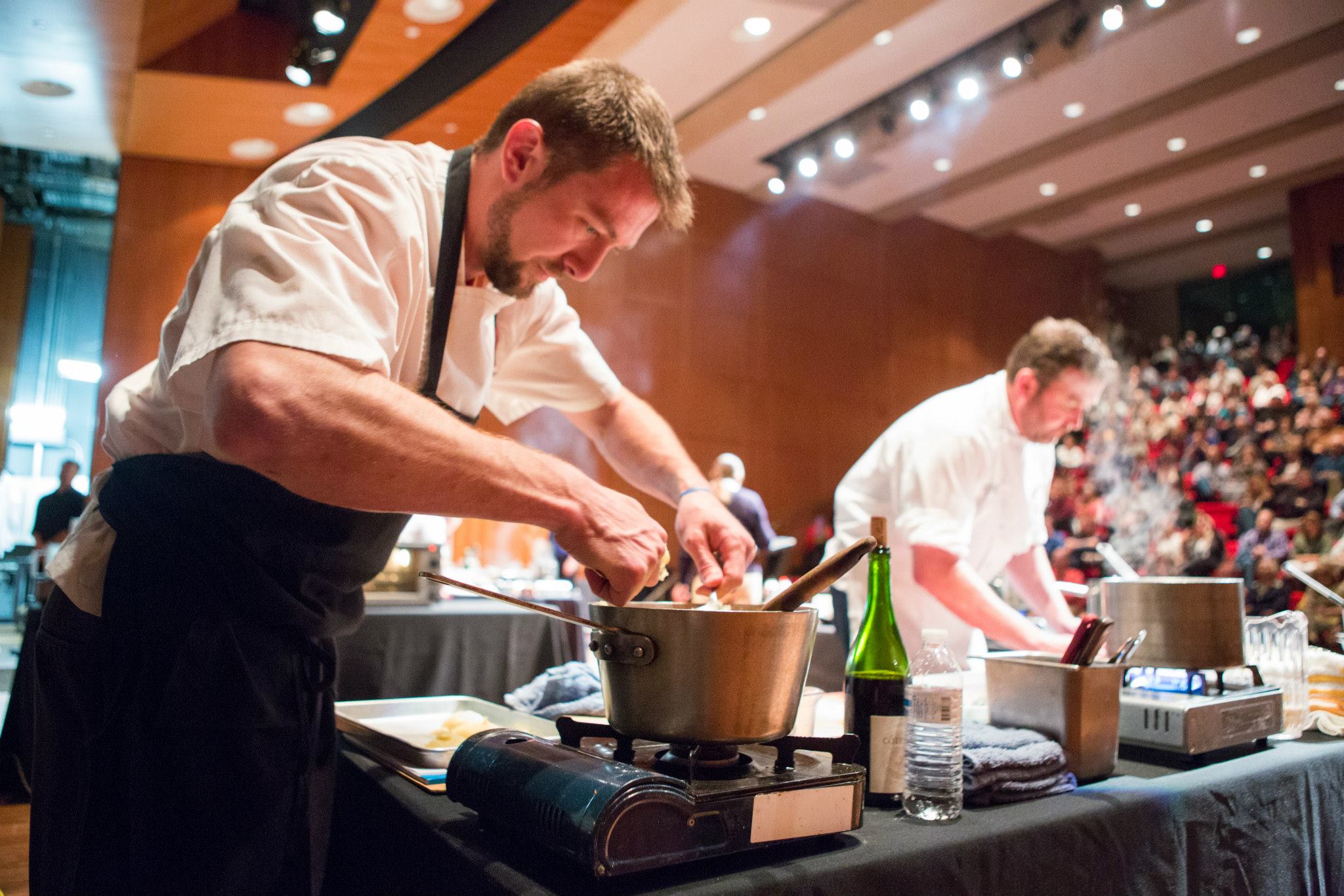 Modern photograph of two white men in chef's uniforms, cooking onstage in the Auditorium in front of a packed audience.