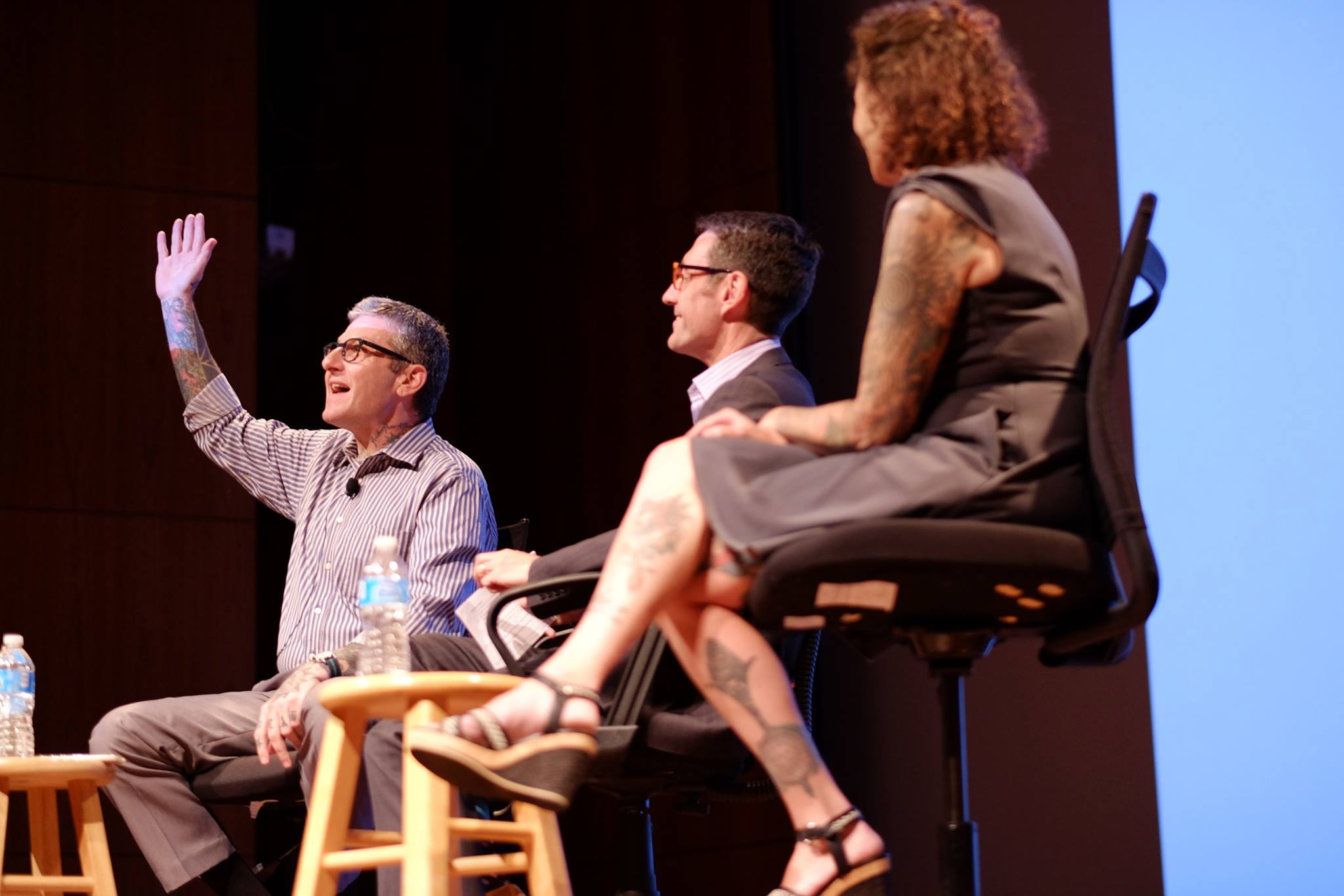 Modern photograph of two older white men and a white woman seated on the Auditorium stage. One white man is speaking to the audience, gesturing with his shirtsleeves rolled back to show his forearm tattoo sleeves. The woman is wearing a knee-length sheath dress that reveals tattoos on her calves.