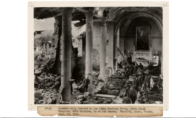 Black and white historical photograph showing the interior of a huge stone building crumbled by bombs. Rows of injured people lie on stretchers inside, tended by nurses and soldiers.