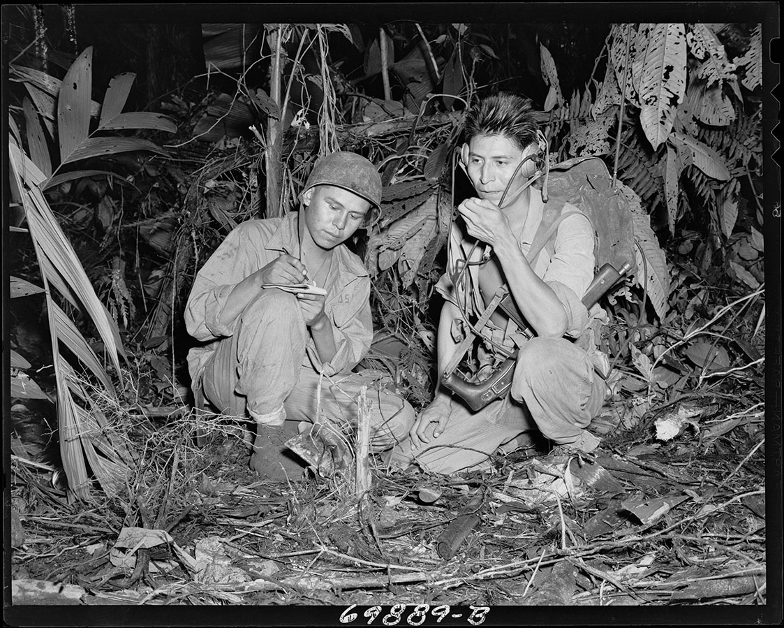 Black and white photograph of two Native American men in WWII combat uniforms kneeling down in a jungle clearing. One is speaking into a portable radio unit while the other is writing on a notepad.