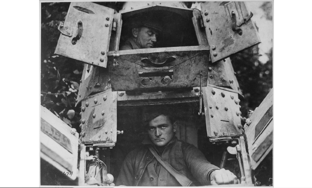 Black and white vintage photograph of a young white WWI soldier sitting in a tank, glaring through the open window into the distance. Another WWI soldier sits above him, visible through another open window in the tank.