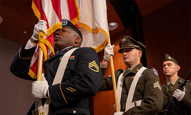 Modern photograph of three members of a color guard dressed in modern U.S. military uniforms, marching in a line. The one in the lead, carrying a U.S. flag, is a Black man.