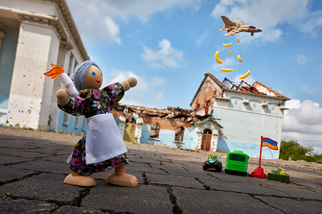 A wooden doll wearing a floral print fress and white apron prepares to throw Lego resembeling a Molotov cocktail. On the ground sits three plastic toy vehicles and a plastic Ukrainian flag. A toy plane flies through the air above, dropping plastic bananas from the sky.