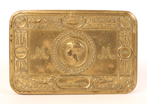 A golden color, metal container with an image of a woman's side profile in the center, "M" to the left and right, and artistic embellishments around the edges.