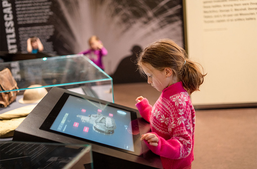 Modern photograph of a small white child in a pink sweater standing at an interactive screen showing a computer-generated model of a WWI tank.