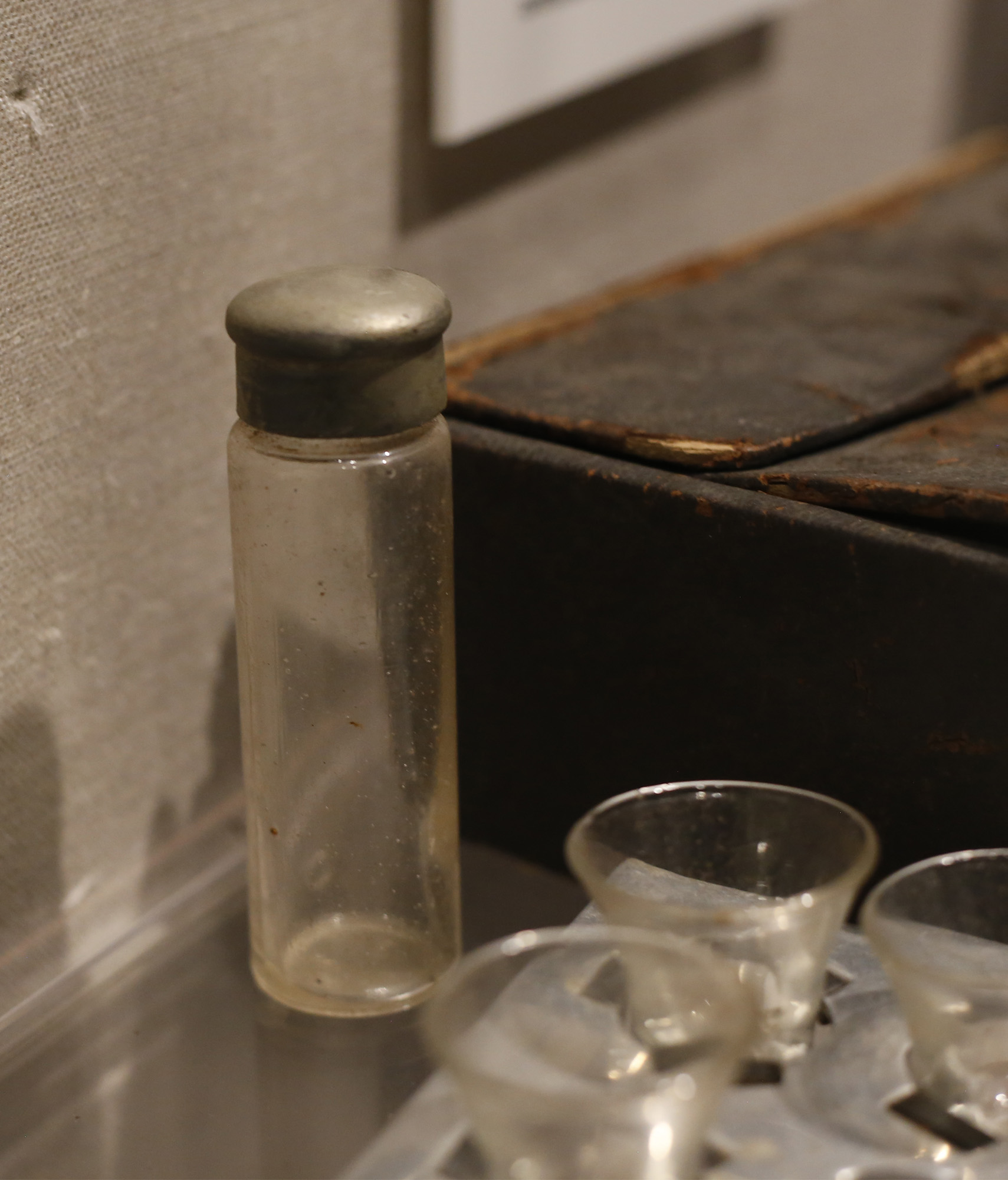 Modern photograph of a small glass vial with a dulled metal cap situated in a museum exhibit case