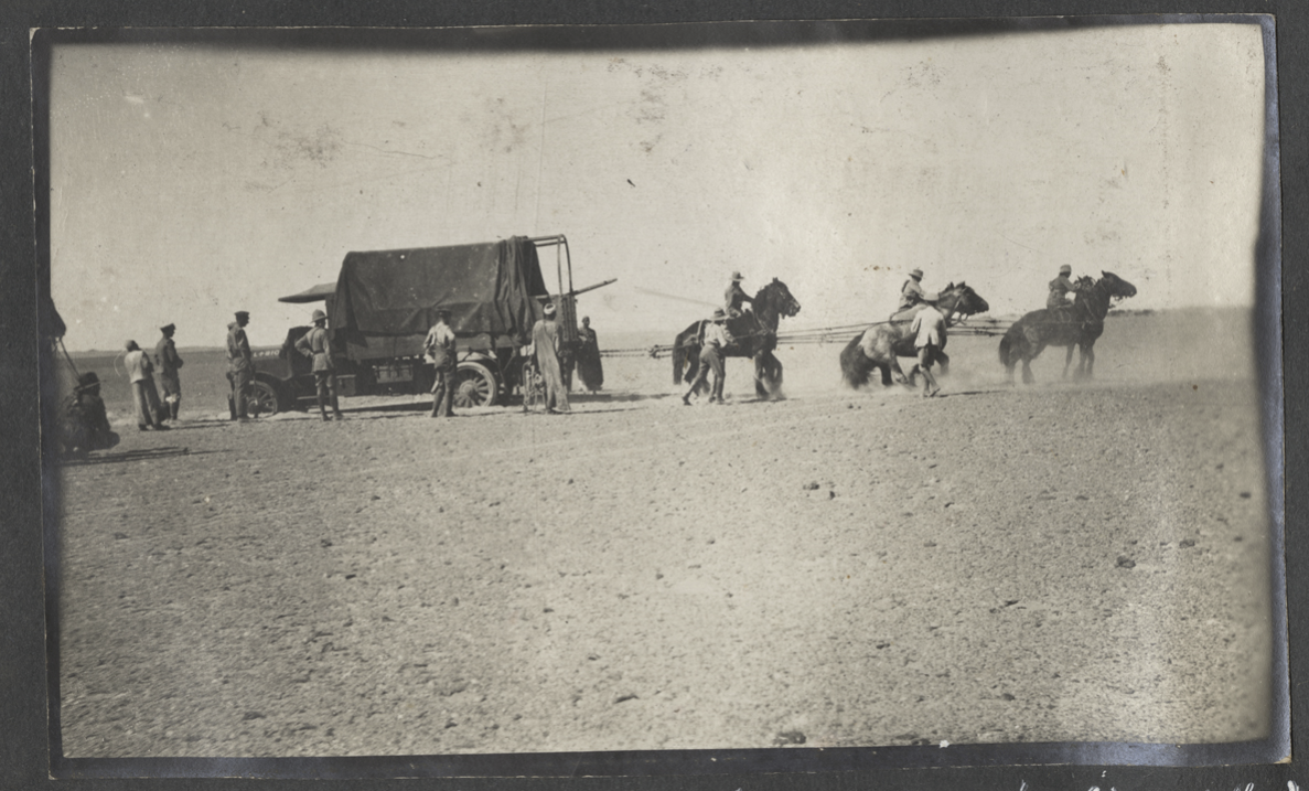 Black and white photo of a WWI-era truck stuck in sand. A team of six horses and their human handlers attempt to pull it out while other soldiers look on.