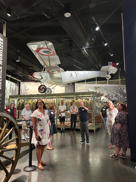 Modern photograph of a history museum gallery dominated by a 3/4 scale model of a WWI-era airplane. Various people grouped under it are looking up at it or at the curator who is speaking.