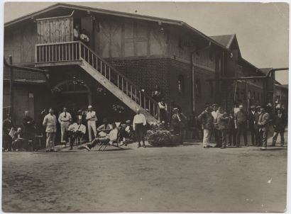 Black and white photograph of inmates at the Ruhleben Internment Camp. Men in a variety of outfits stand along the ground in front of a two-story building, while others stand on the second floor.