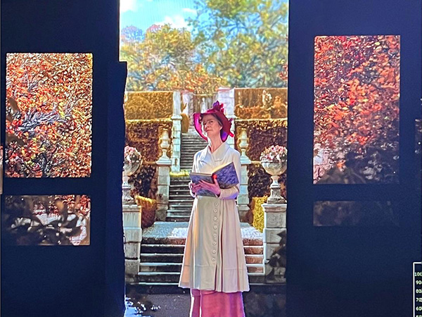 Screenshot of a white female actor in a WWI-era dress and red hat composited onto a CGI background of a European garden