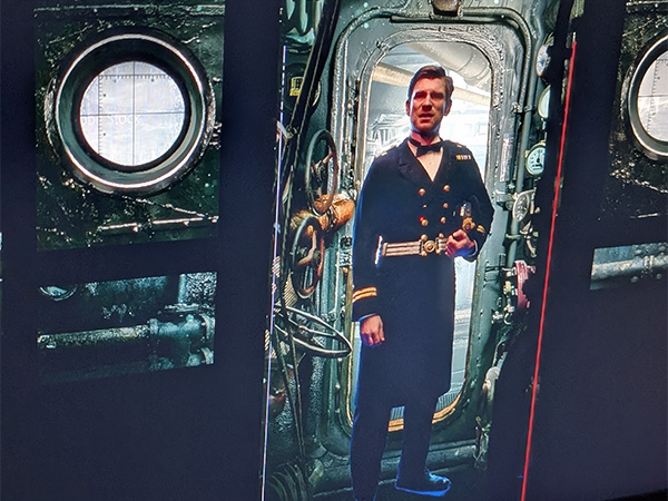 Modern photo of a computer monitor showing a white male actor in a WWI military uniform composited onto a CGI background of the inside of a submarine