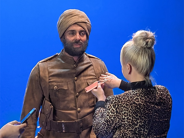 Modern photo of a brown-skinned, bearded male actor in a WWI military uniform and turban, standing in a bluescreened studio while a wardrobe department person adjusts his costume