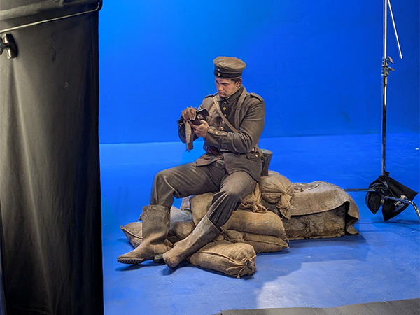 Modern photo of a white male actor in WWI military uniform sitting on a pile of sandbags in a bluescreened studio, surrounded by lights and reflector panels