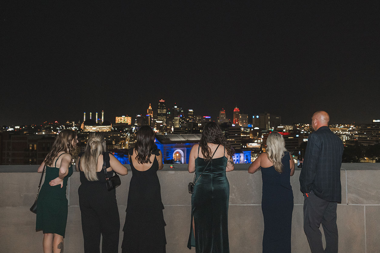 Photo of a group of partygoers with their backs to the camera, looking out to the nighttime Kansas City skyline