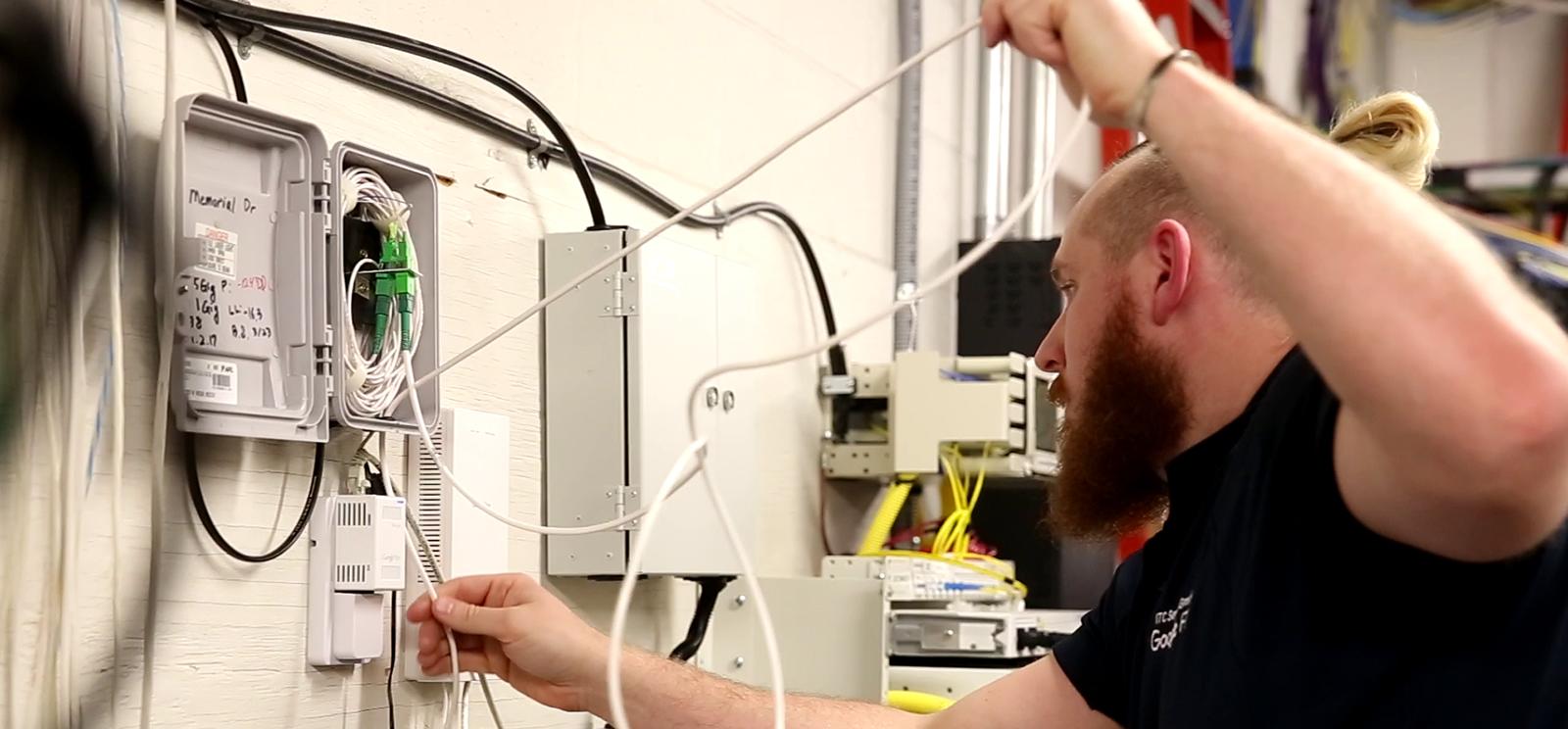 Modern photograph of a bearded white man threading cables through a complicated tangle of cables installed into a wall.