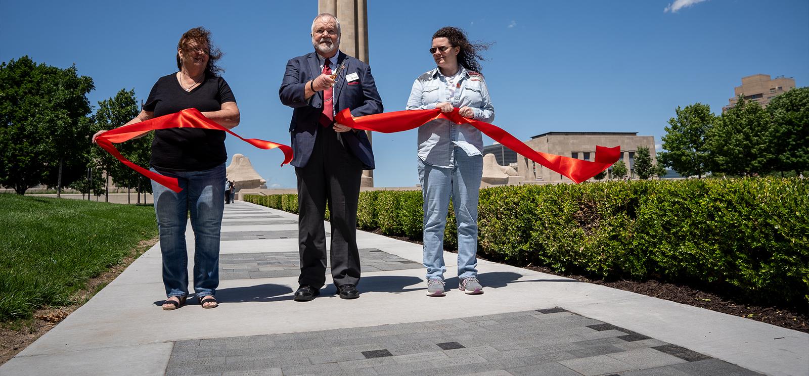 Modern photograph of the west sidewalk looking toward Liberty Memorial Tower, which has hundreds of dark granite bricks inlaid in it. An older white man in a suit ceremonially cuts a red ribbon that two white women are holding between them.