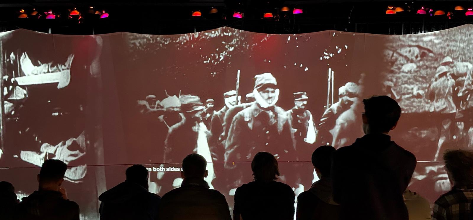 Modern photograph of museum guests, turned away from the viewer, silhouetted by a large movie screen that is projecting black and white images of WWI soldiers