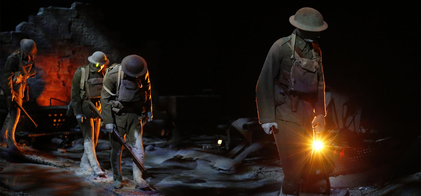 Modern photograph of life-size WWI soldier mannequins trudging across a muddy battlescape, lit starkly by orange and white spotlights