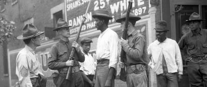 Black and white photograph of three white men in National Guard uniform holding rifles surrounding a Black man in civilian clothing. Two Black men in civilian clothing observe off to the side. A Black man in military uniform also observes.