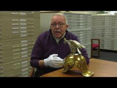 Video still of an older white man wearing glasses and white gloves seated at a table in a room filled with metal cabinets. On the table in front of him is a shiny, engraved metal helmet.