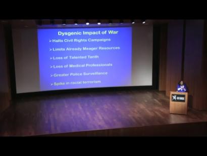 Video still of a Black woman at a podium on the Museum auditorium stage giving a presentation.