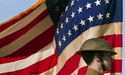 Modern photograph of a man in WWI-era uniform, visible only from the neck up, framed by an enormous U.S. flag blowing in the wind.