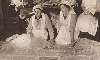 Black and white photograph of two white women in white pinafores kneeling on the ground surrounded by newspapers